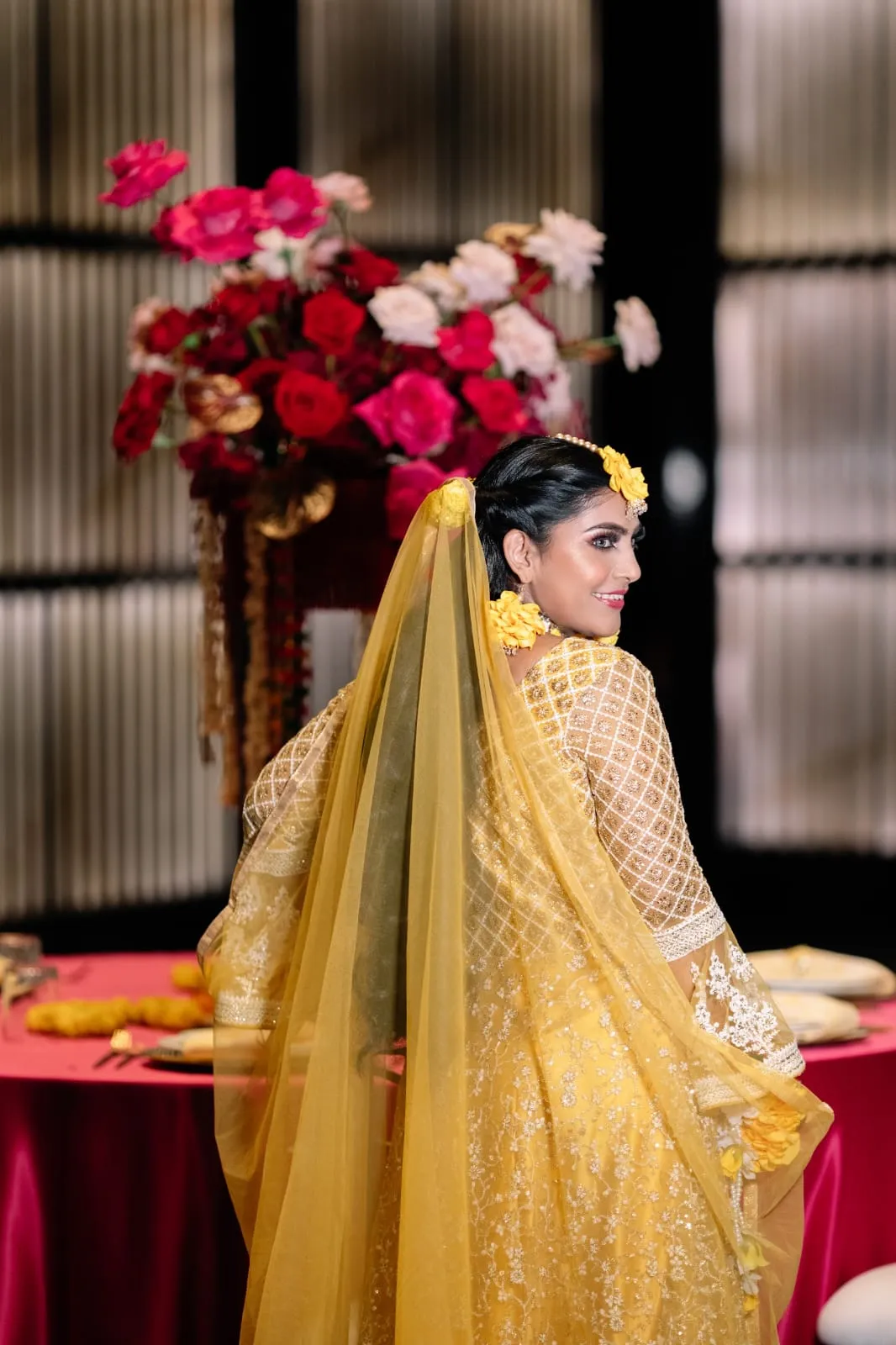 Woman in a gold-yellow outfit and veil seen from behind, seated in front of red flowers