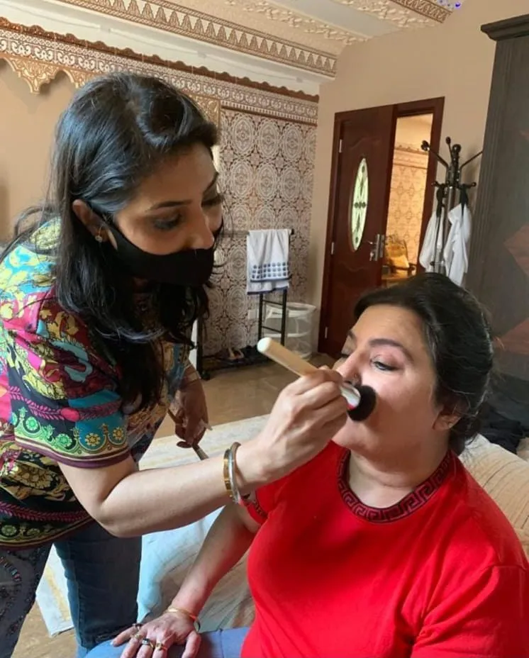 Makeup artist in a colourful jacket and mask applying powder to a woman in a red top seated at a window