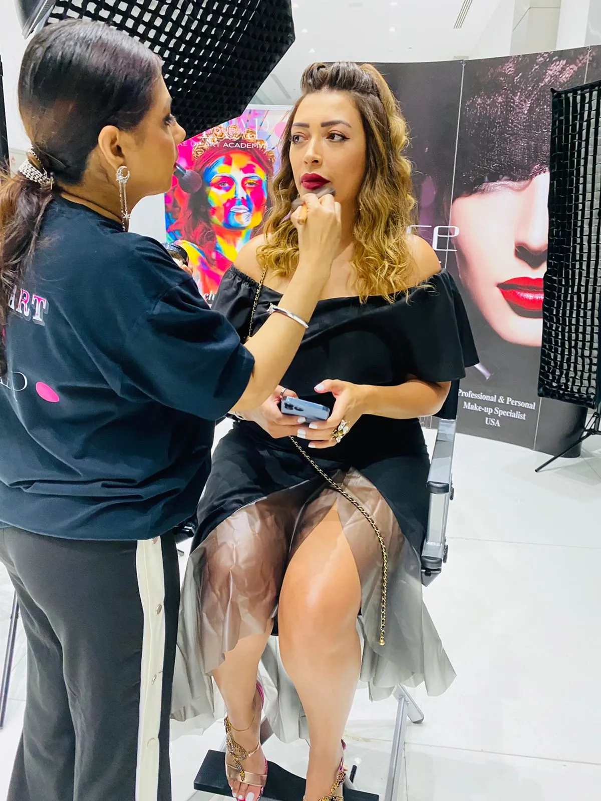 Makeup artist in a navy top working on a woman in a black ruffled outfit at a beauty stand