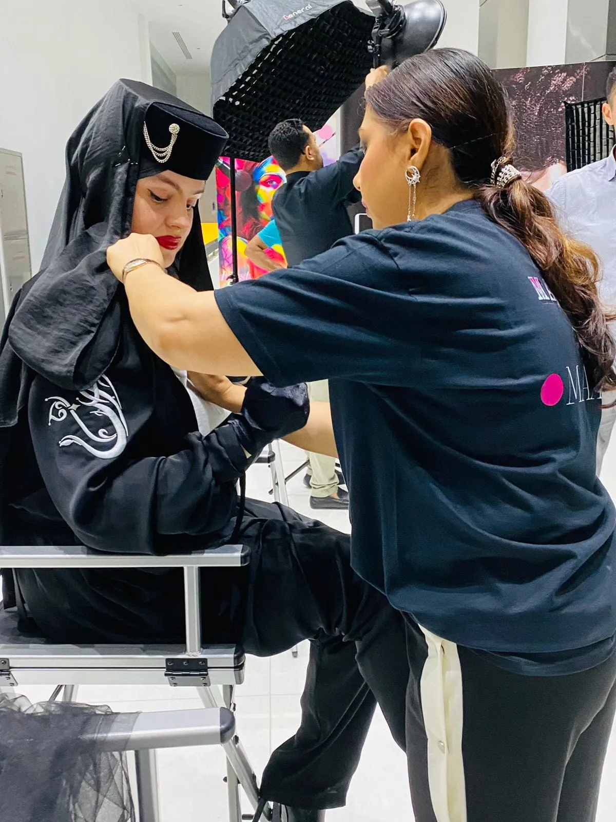 Makeup artist in a navy t-shirt working on a person in a black hooded costume, seen from another angle