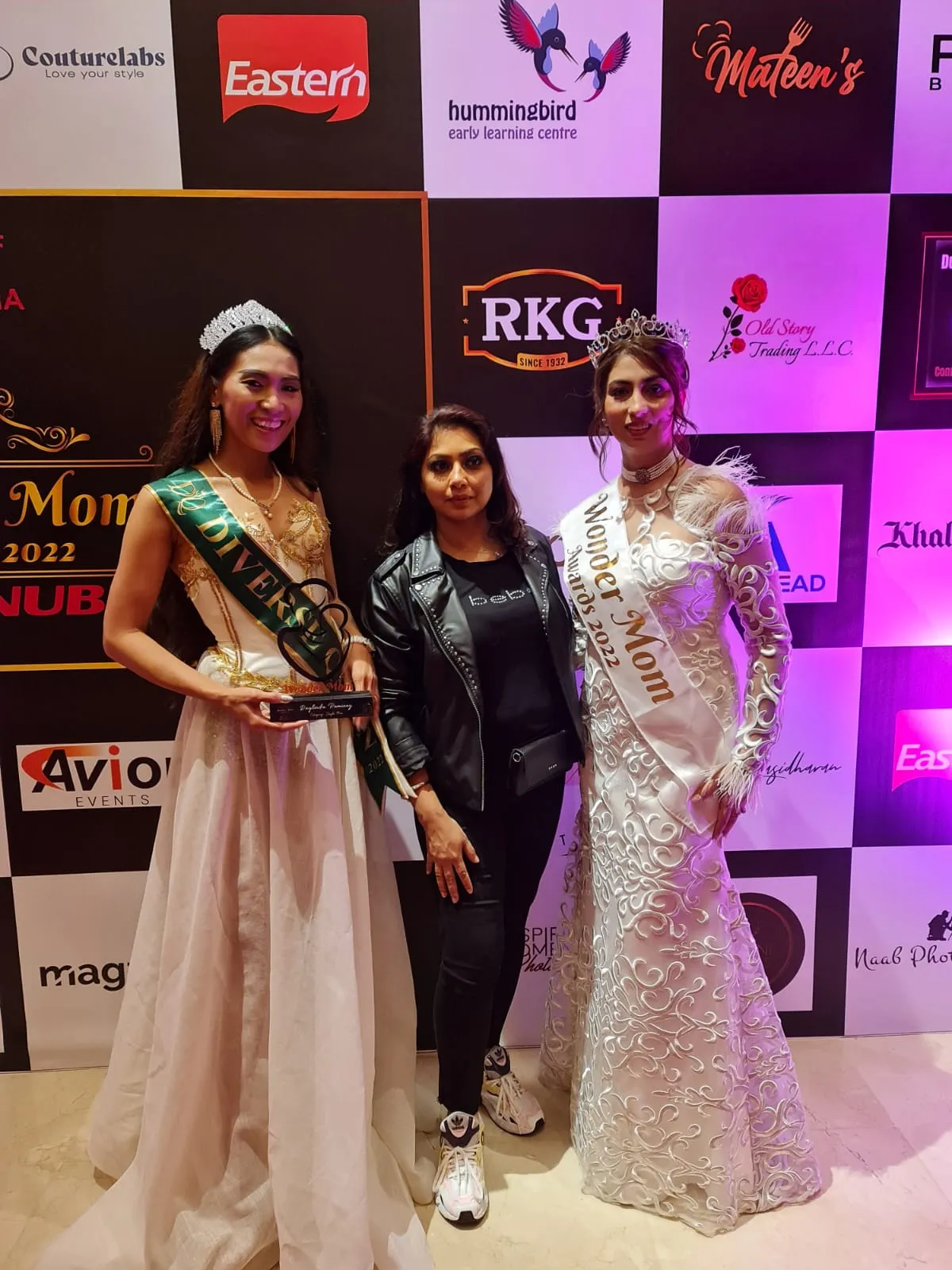 Two women in long gowns with sashes and tiaras, and a woman in black between them, in front of a sponsor backdrop