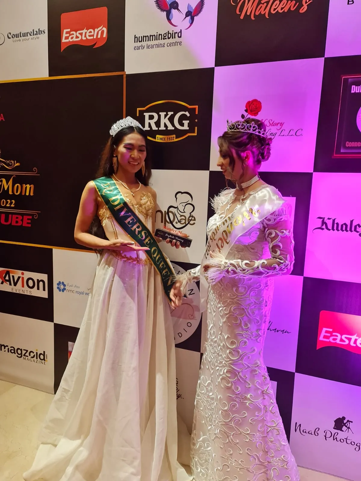 Two women in long gowns with sashes and tiaras photographed from a closer angle in front of a sponsor backdrop