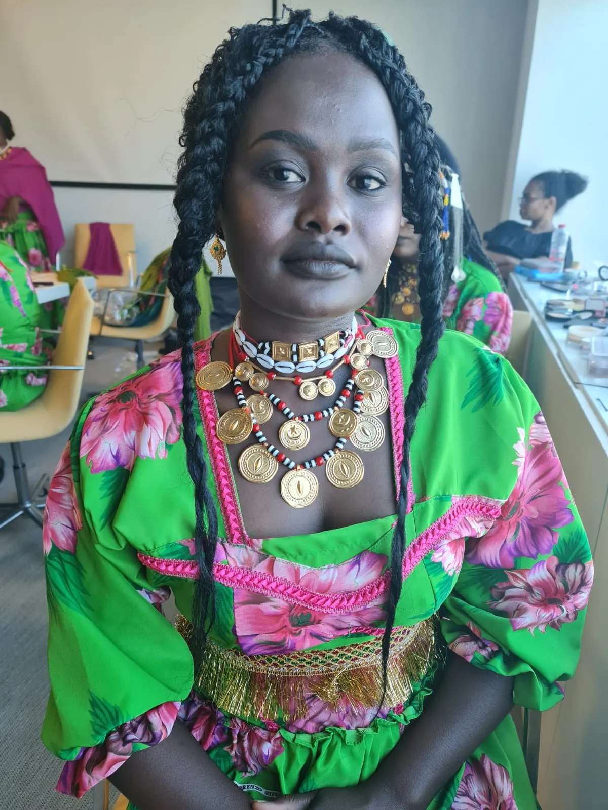 Woman with braided hair in a bright green and pink patterned outfit and chunky gold necklaces
