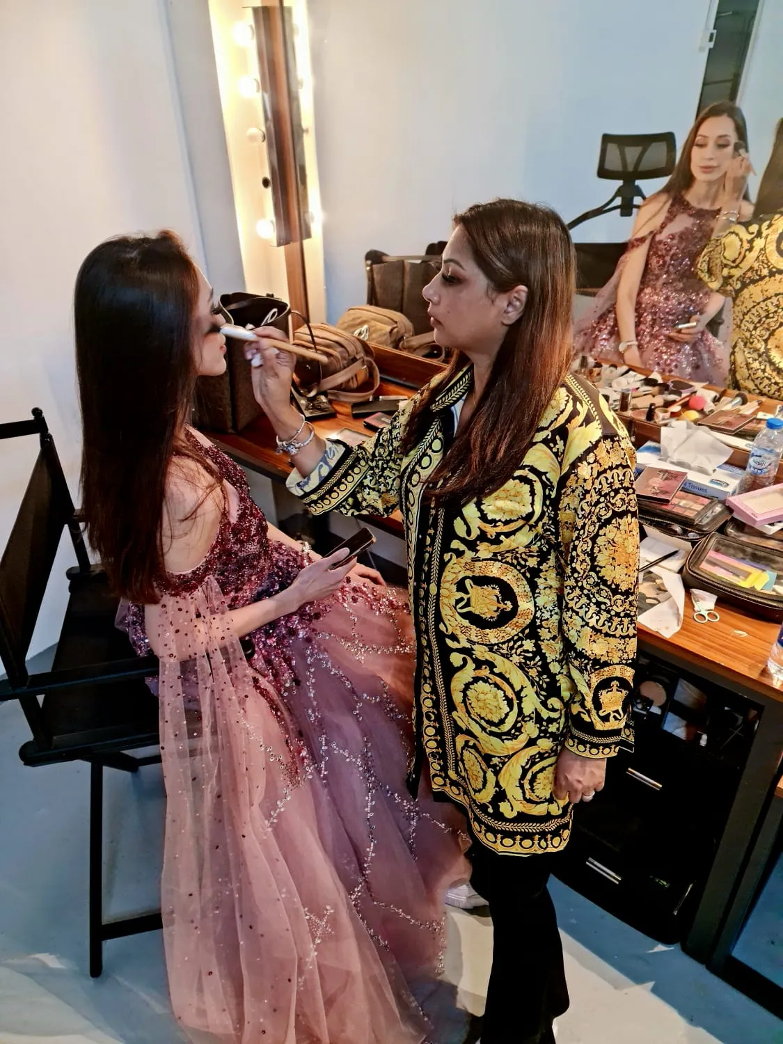 Makeup artist in a black and gold jacket applying makeup to a woman in a pink dress