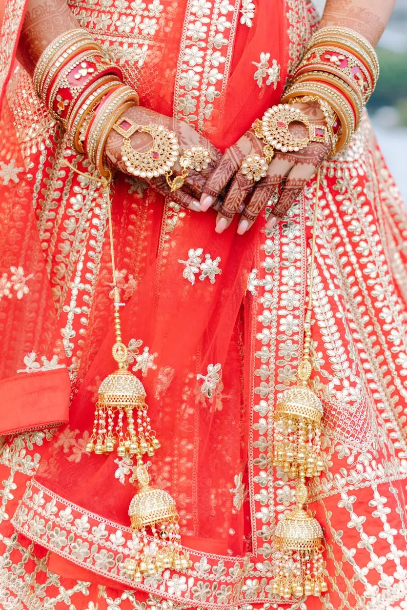 Hands wearing red bangles and rings holding gold bridal ornaments over a red embroidered outfit