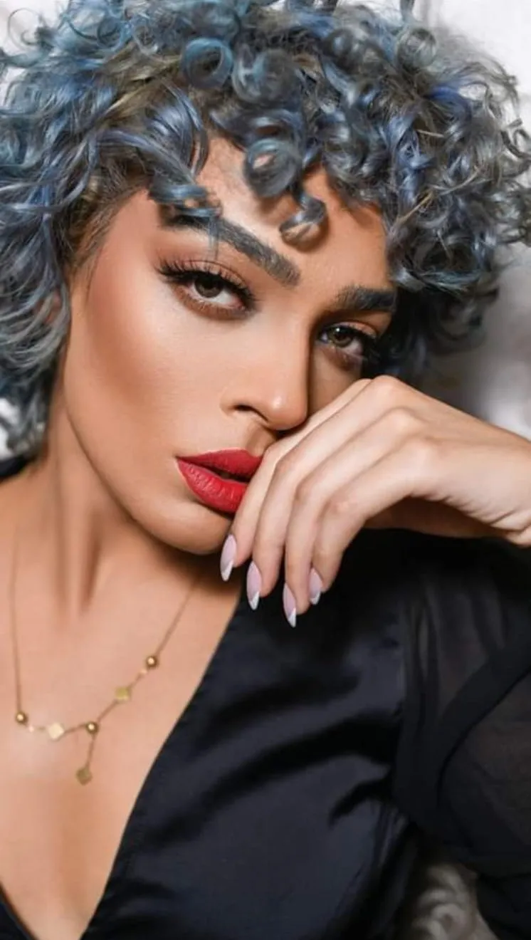 Close-up of a woman with silver-blue curly hair and bold red lips, one hand under her chin