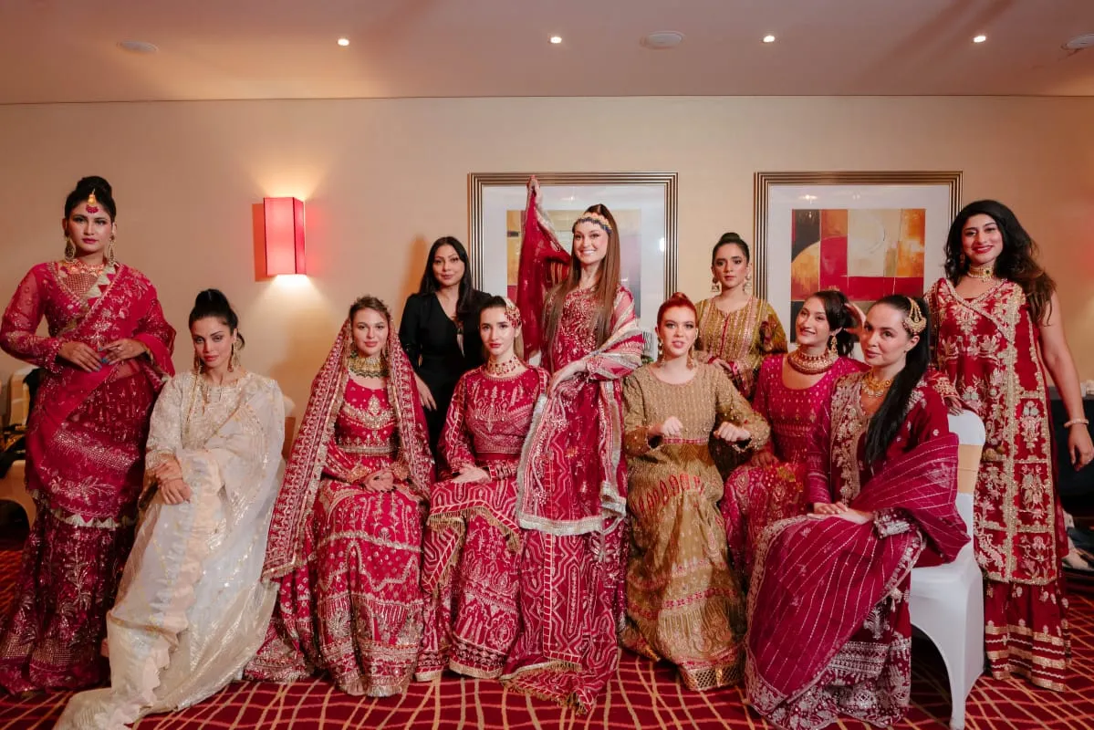 Group of women in red and gold outfits gathered around a woman in ivory in a hotel room