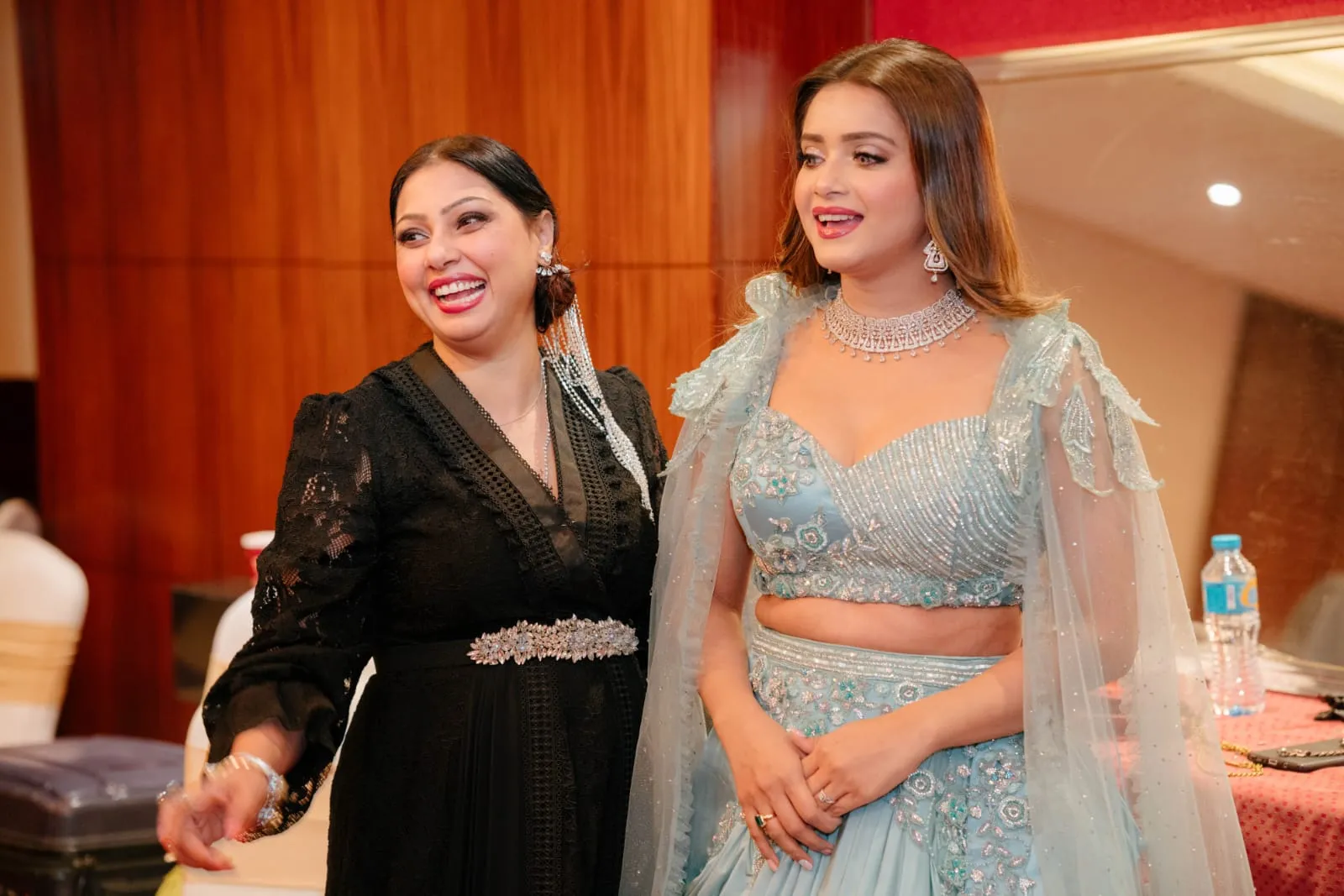 Two women posing together in a room with orange walls, one in black and one in a pale blue embellished outfit