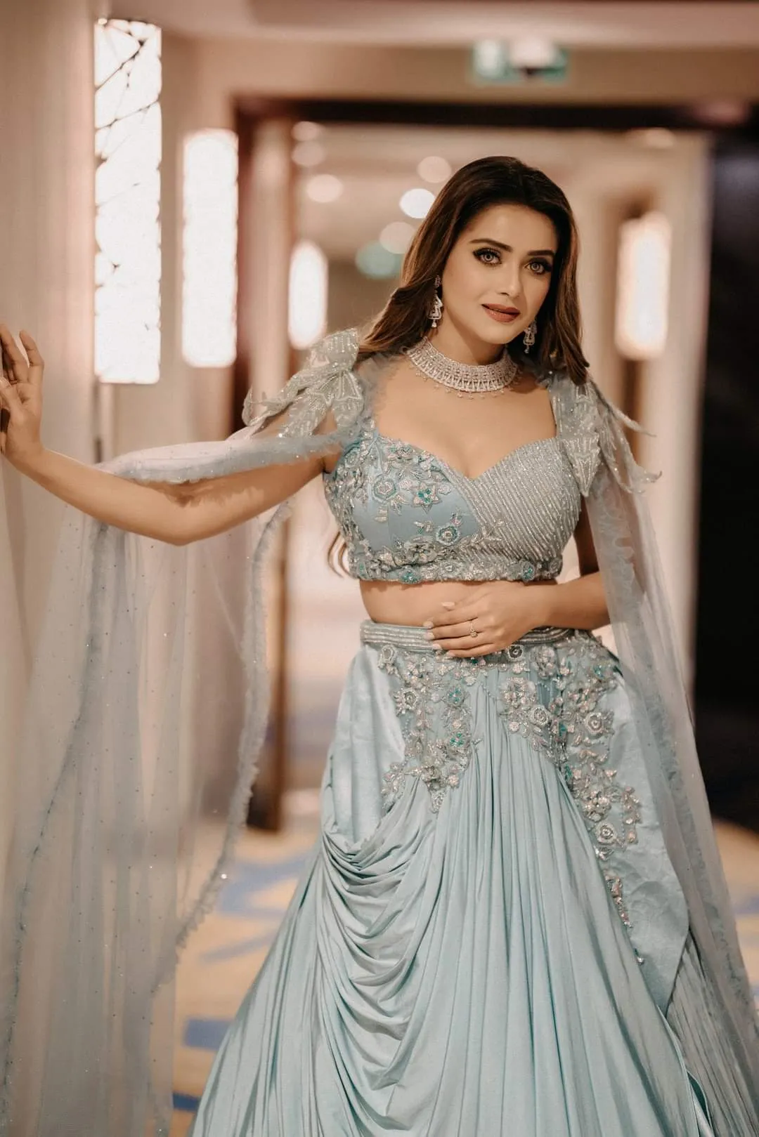 Woman in an embellished sage-green gown with one arm stretched out to the side in a hotel corridor