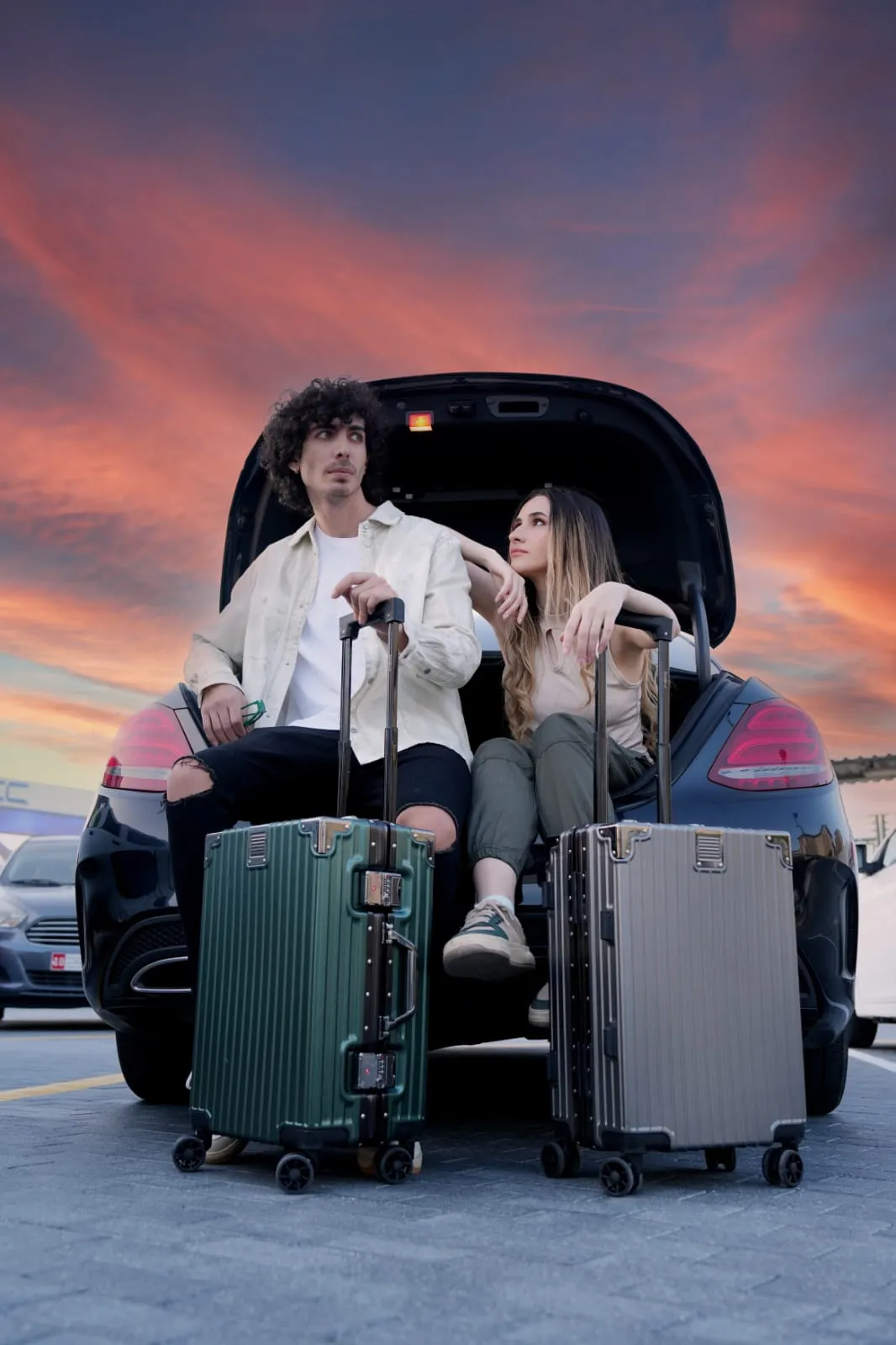 Two people sitting in the open boot of a car at sunset with three suitcases