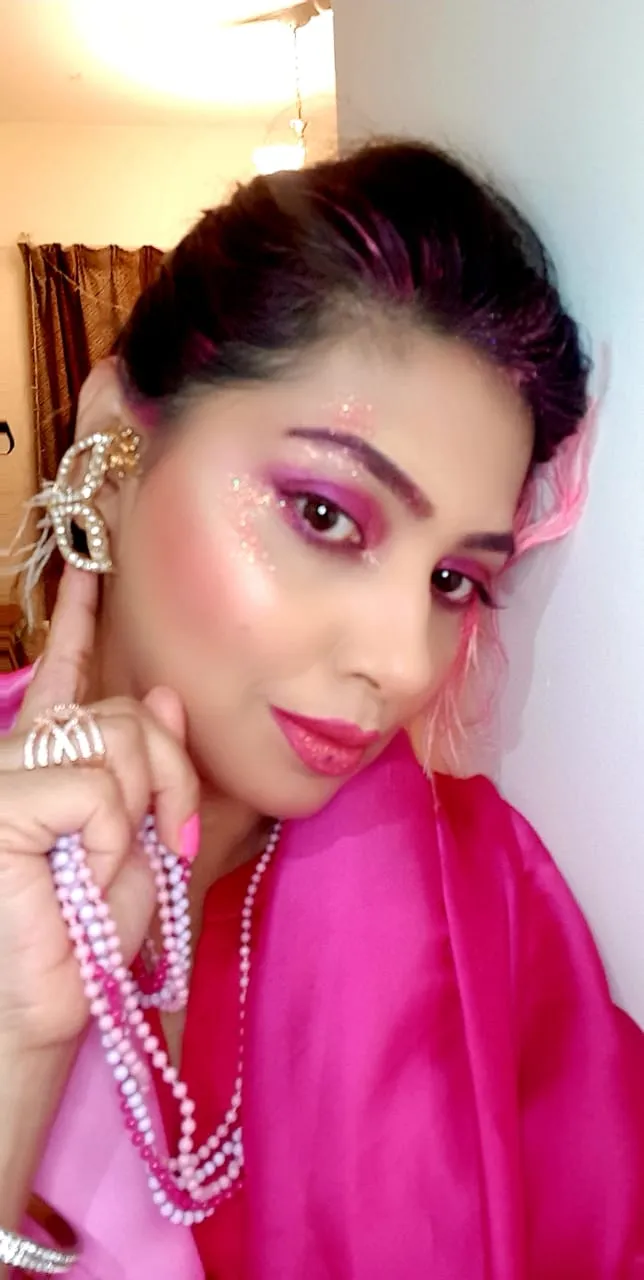 Woman with pink and purple eye makeup in a pink dupatta and pearl necklace, one hand at her earring