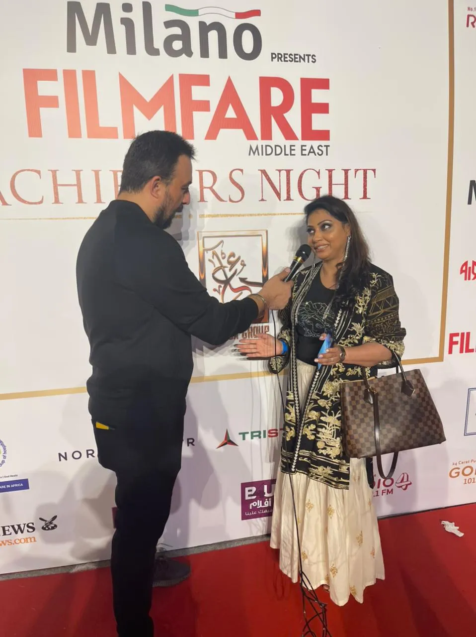 Man speaking with a woman in a black and gold jacket in front of an awards-night backdrop