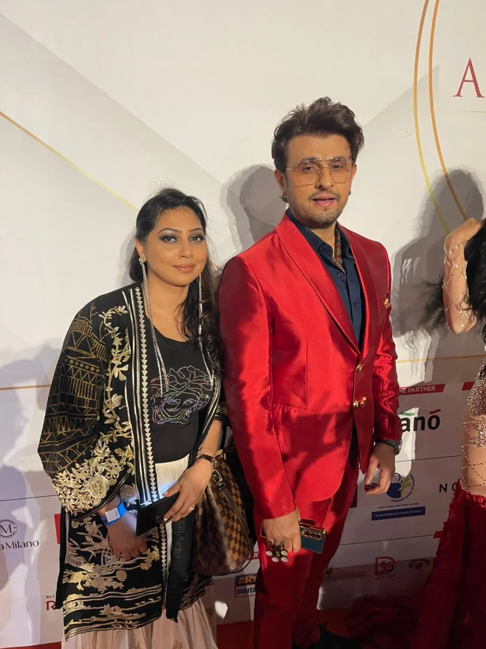 Woman in a black and gold jacket posing with a man in a bright red suit in front of an awards-night backdrop