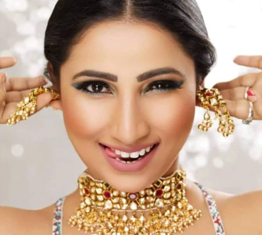 Smiling woman in a gold choker and statement earrings with bright makeup and a hand at each ear