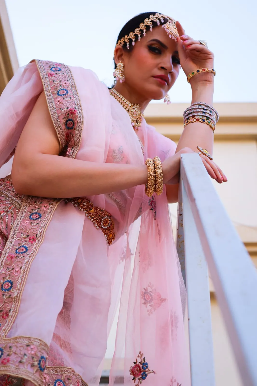Woman in a pink embroidered outfit with gold jewellery leaning on a white railing
