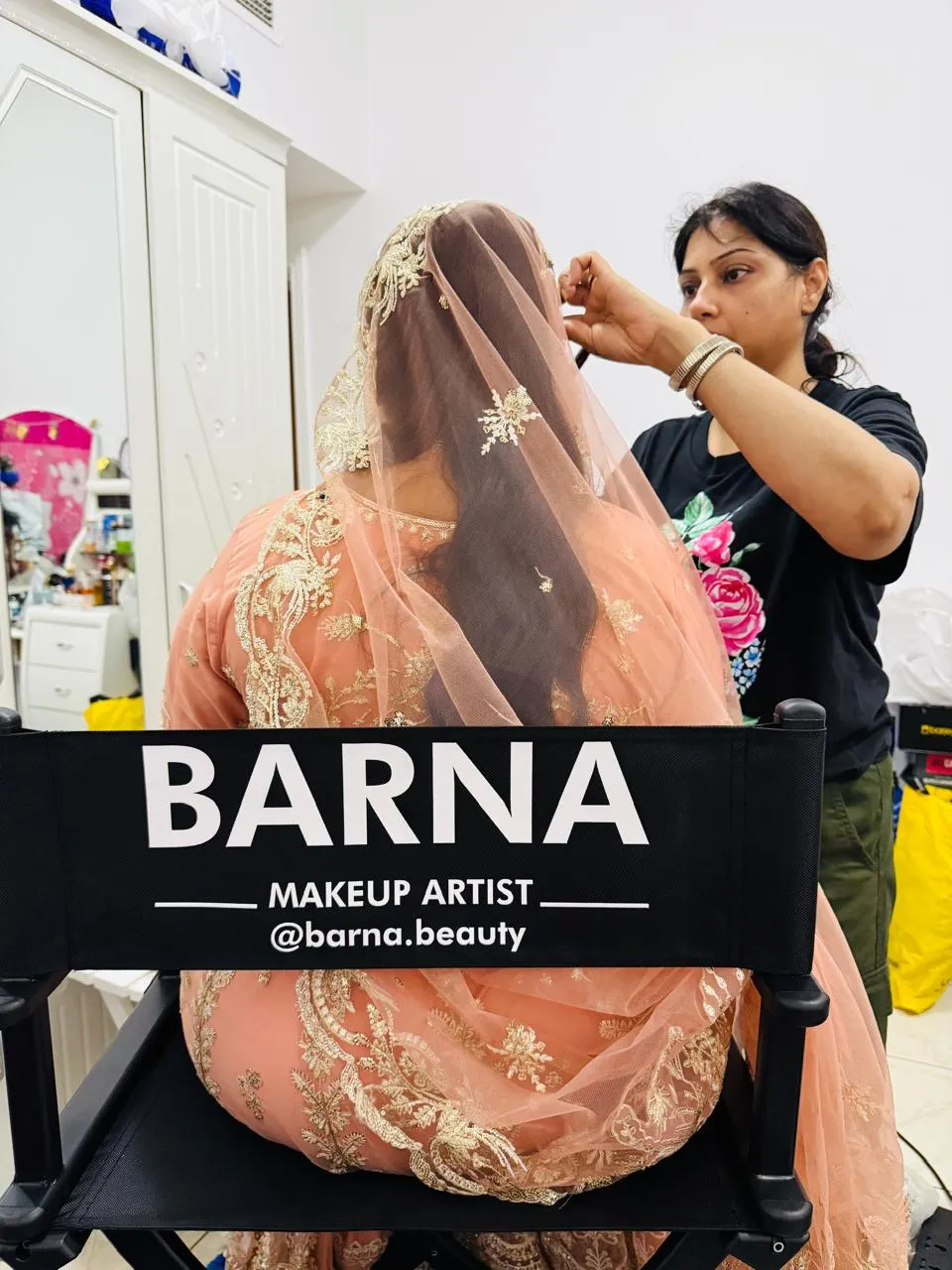 Makeup artist setting the embroidered peach veil of a bride seated in a chair marked Barna makeup artist