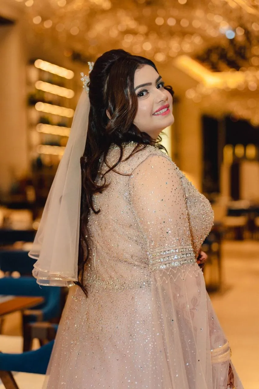 Bride in a blush embellished gown and short veil smiling back over her shoulder in a room with golden lights