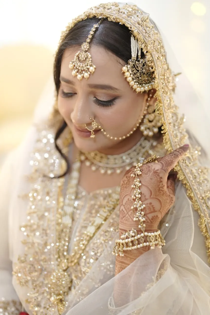 Bride in an ivory and gold embroidered dupatta with a gold maang tikka, nose ring and hand jewellery, eyes lowered
