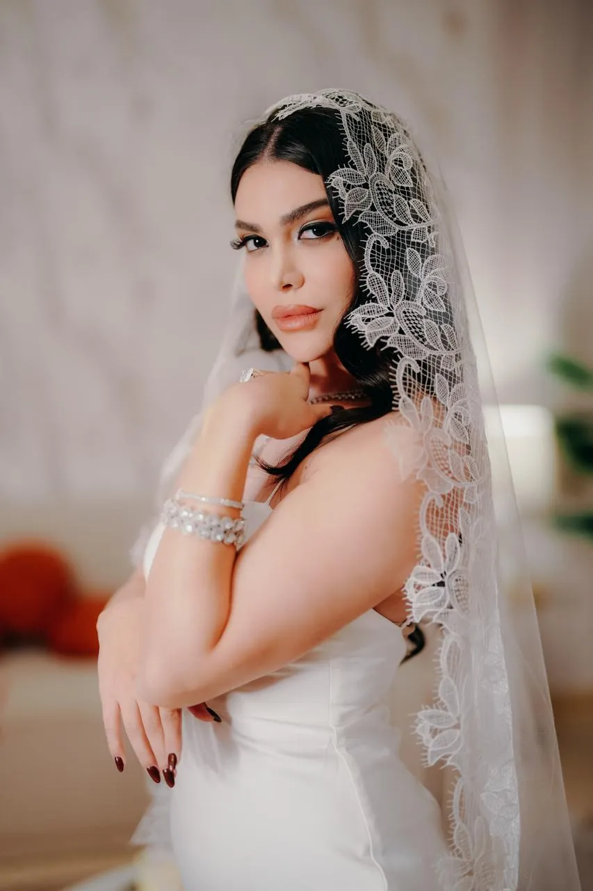 Bride with dark hair in a white gown and a long lace-trimmed veil, arms crossed in front of her