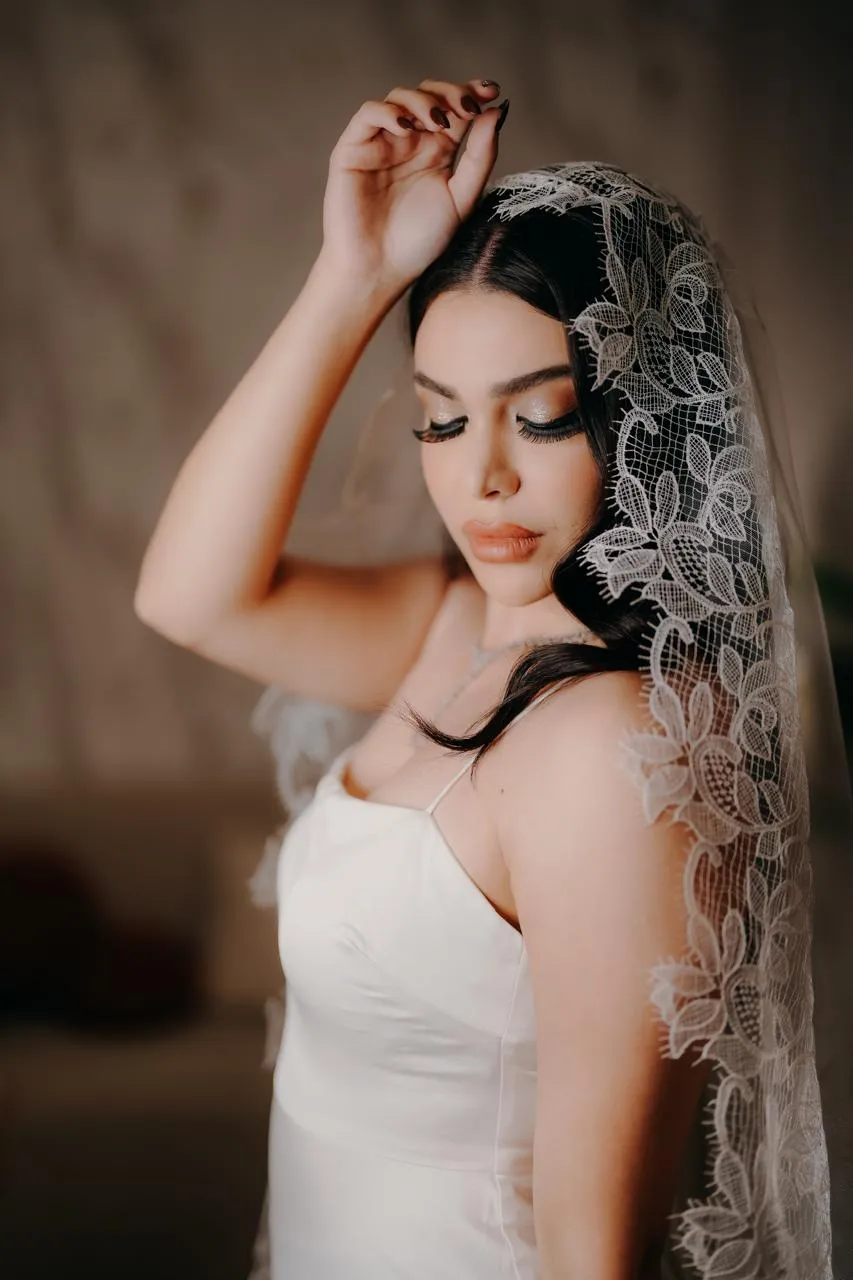 Bride in a strapless white gown holding up the edge of her lace veil with one hand