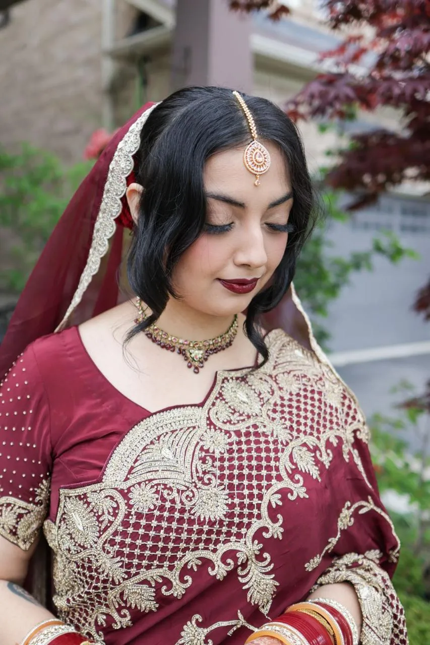 Bride in a maroon and gold embroidered outfit and dupatta with a maang tikka and gold choker, eyes lowered