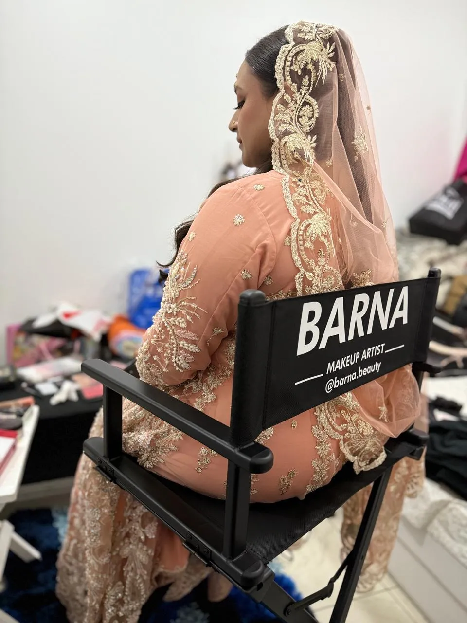 Bride in a peach lehenga and embroidered dupatta seen from behind, seated in a chair marked Barna makeup artist