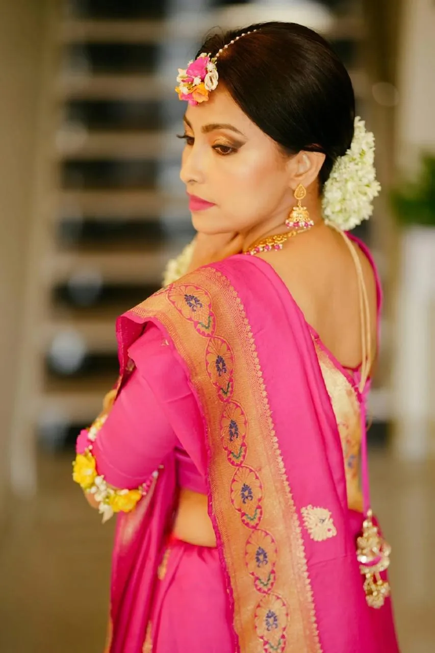 Bride in a pink and gold silk saree with a floral maang tikka and flower bracelets, seated on a lawn and smiling