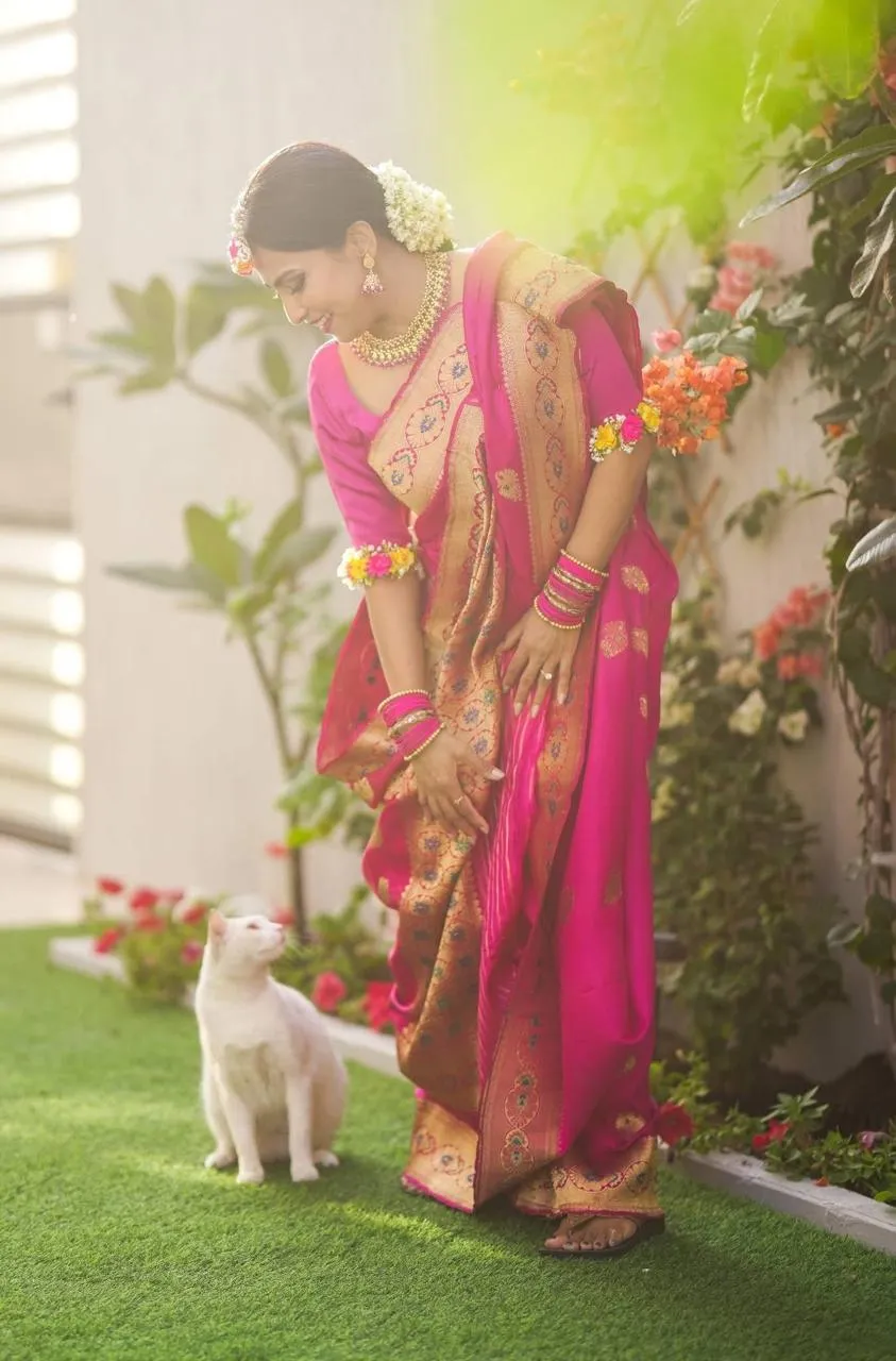 Bride in a pink saree and flower jewellery bending towards a white cat on a garden lawn, full length