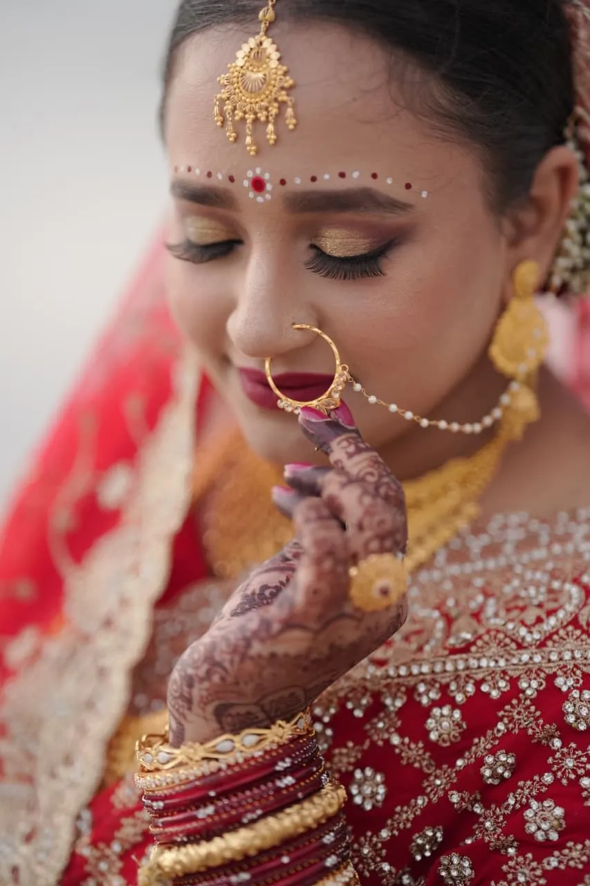 Bride in a red and gold lehenga with a nose ring and maang tikka, a mehndi-painted hand at her chin