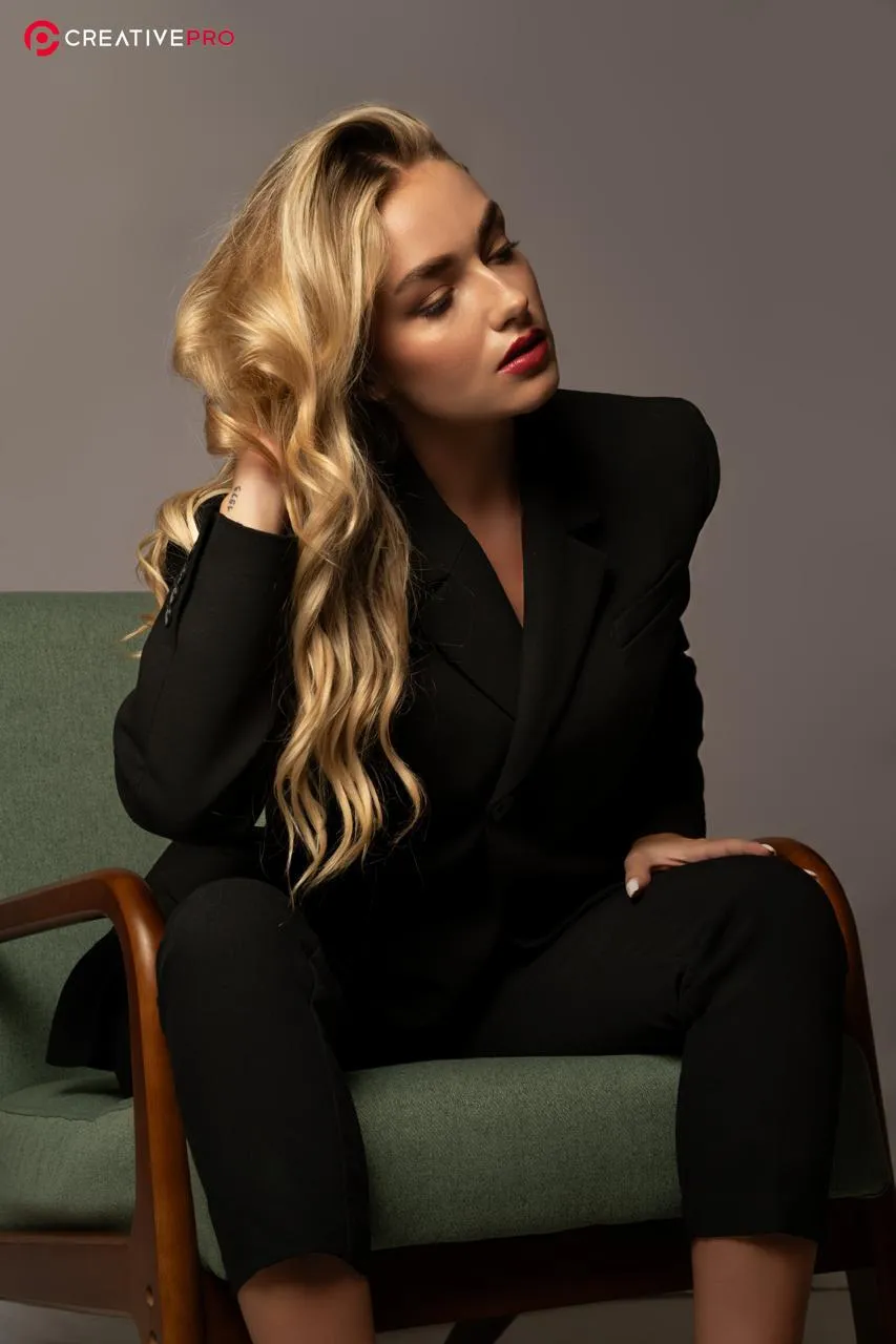 Woman with long blonde waves in a black trouser suit seated in a green armchair