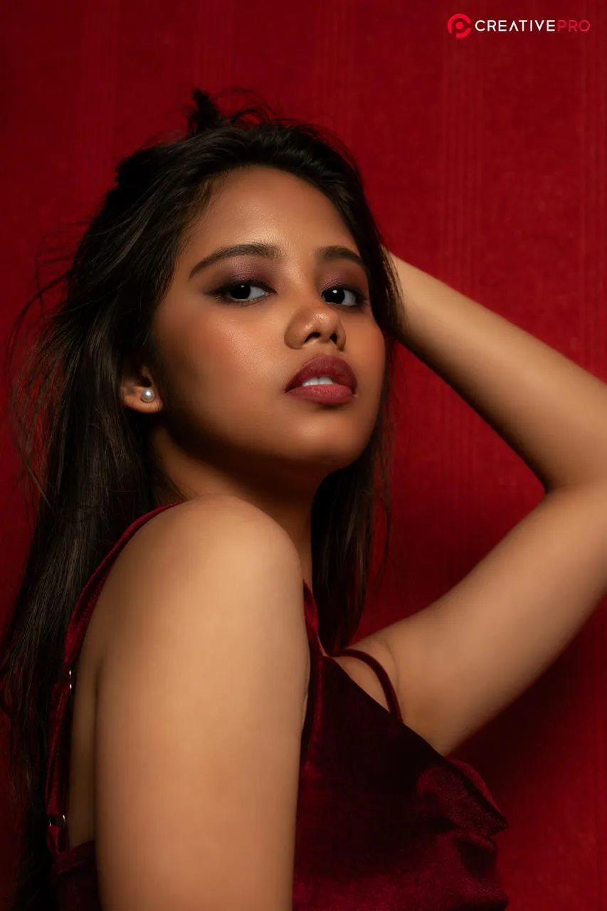 Woman with dark hair and a deep red lip posing with one arm raised against a red backdrop
