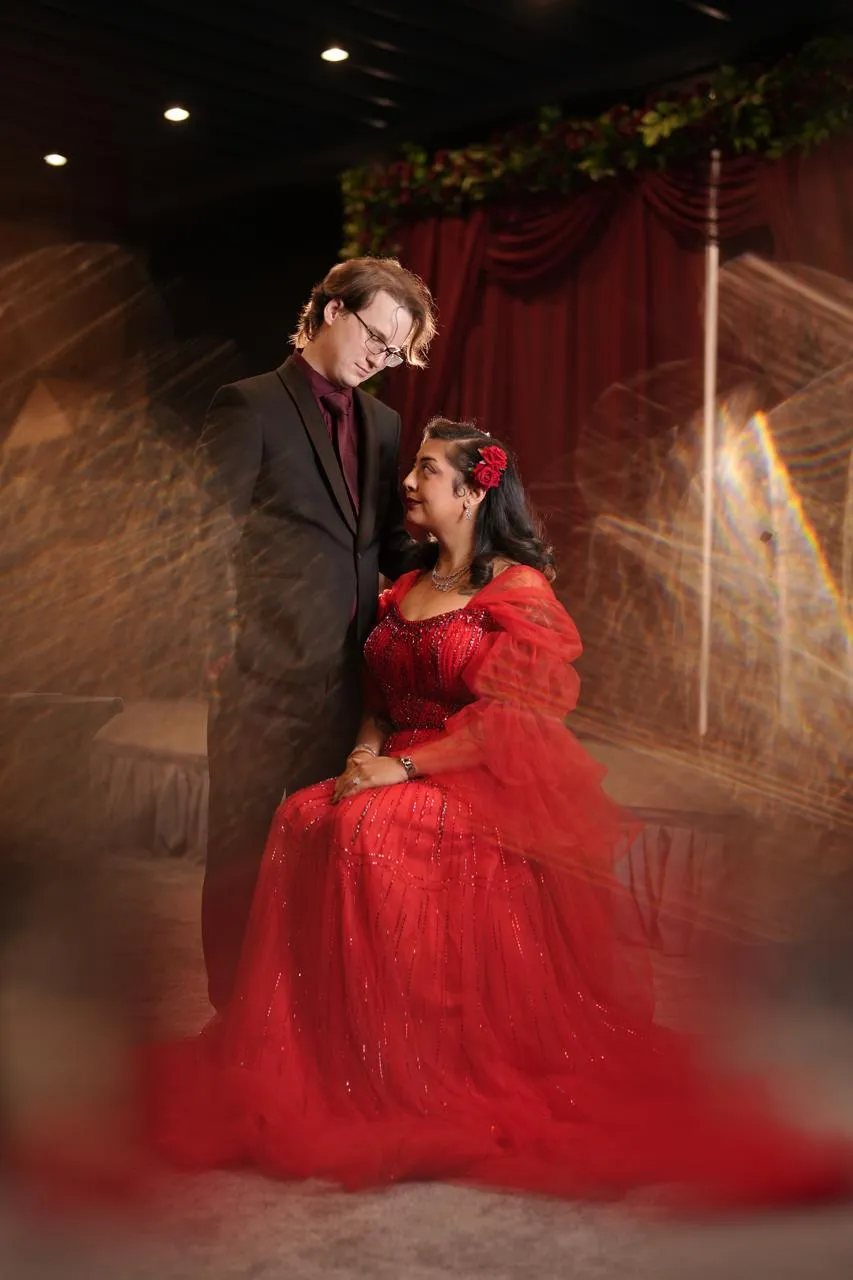 Woman in a red tulle gown seated beside a man in a dark suit in front of red stage curtains