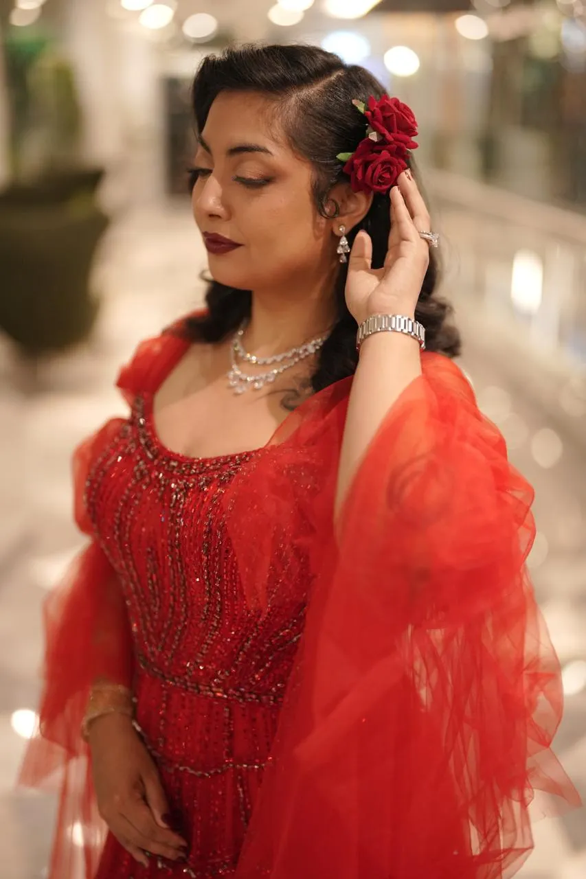 Woman in a red tulle gown with a red rose in her hair and a pearl necklace, a hand at her ear