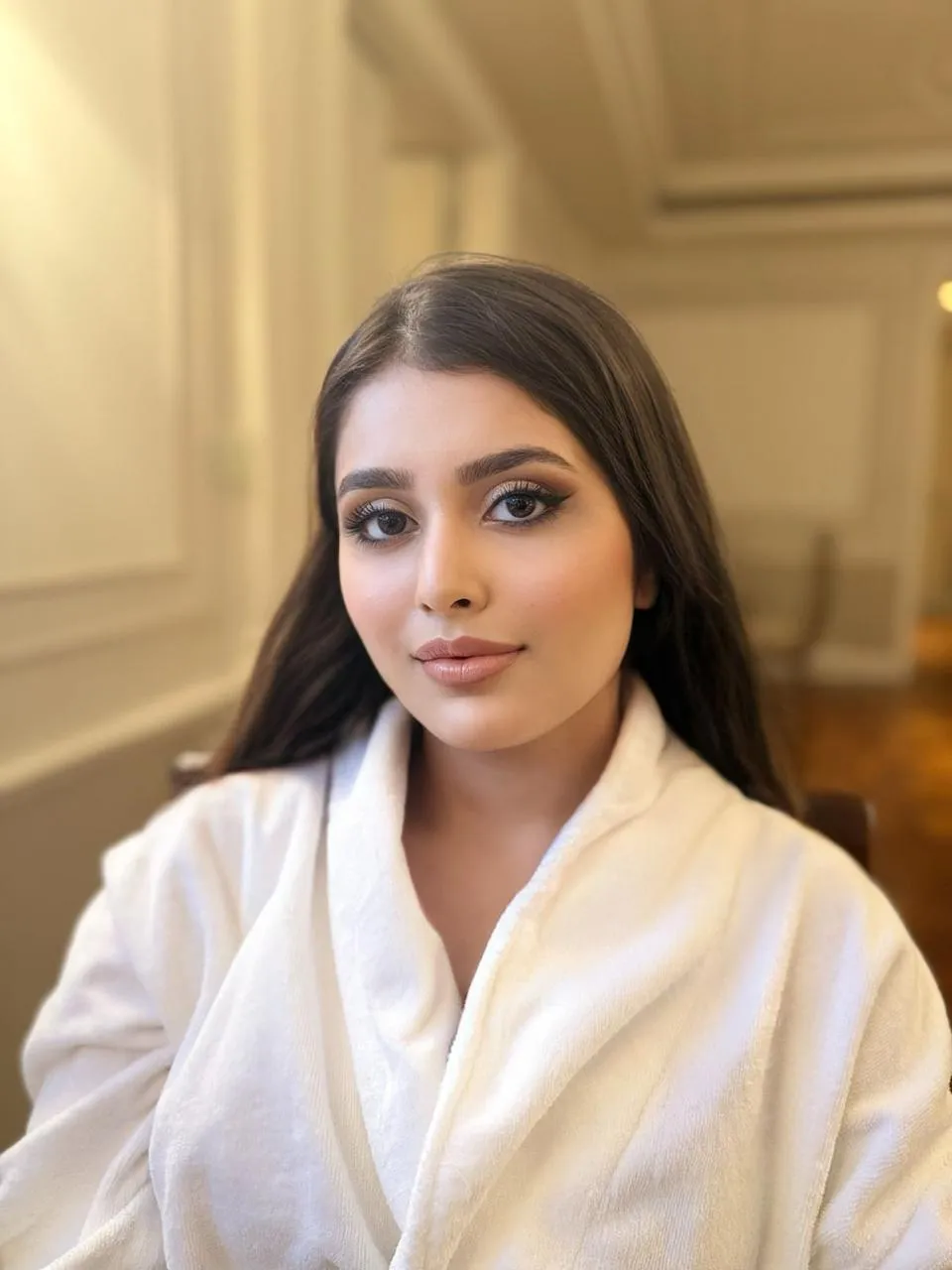 Woman with long dark hair and soft glam makeup in a white bathrobe, seated in a room with panelled walls