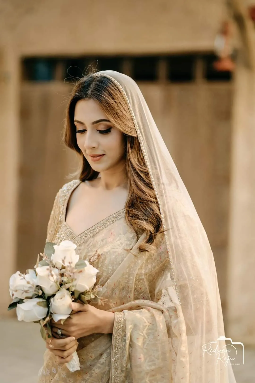 Bride in an ivory and gold outfit with a sheer veil, holding a bouquet of white roses