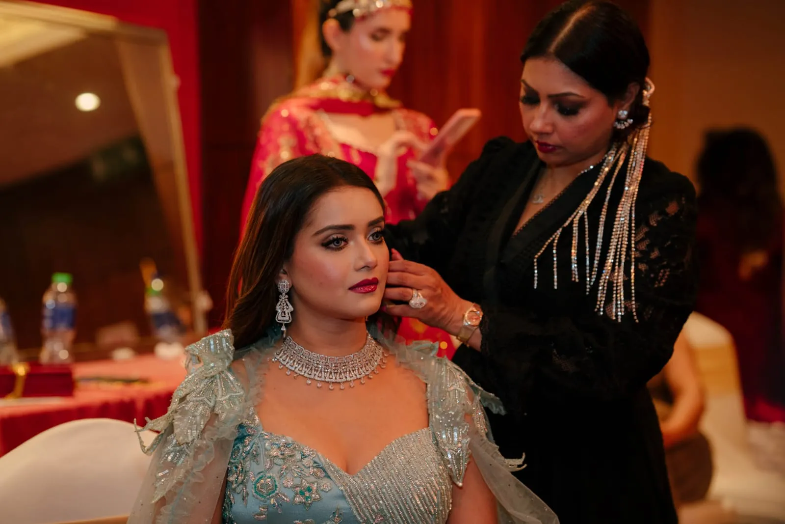 Makeup artist in black styling the hair of a model in a sage-green embellished outfit and layered necklace, backstage at an event