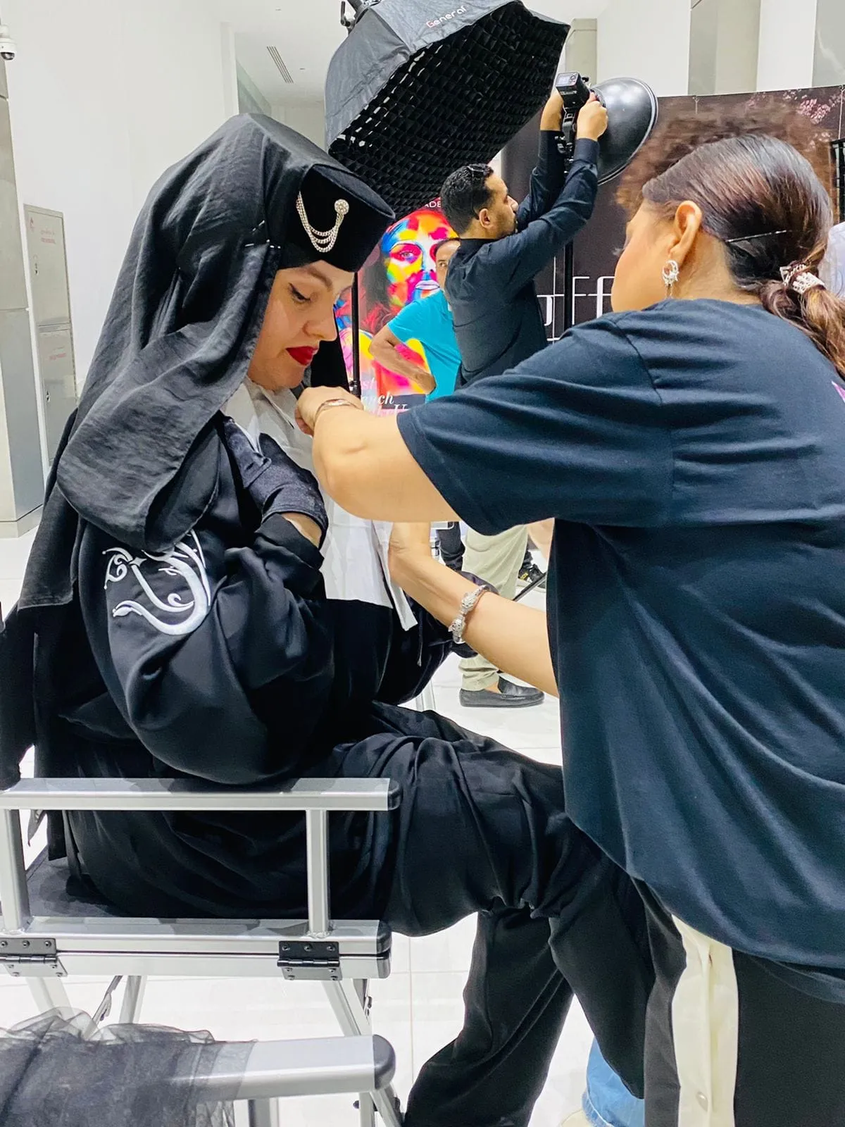 Makeup artist in a navy t-shirt working on a seated person in a black hooded costume