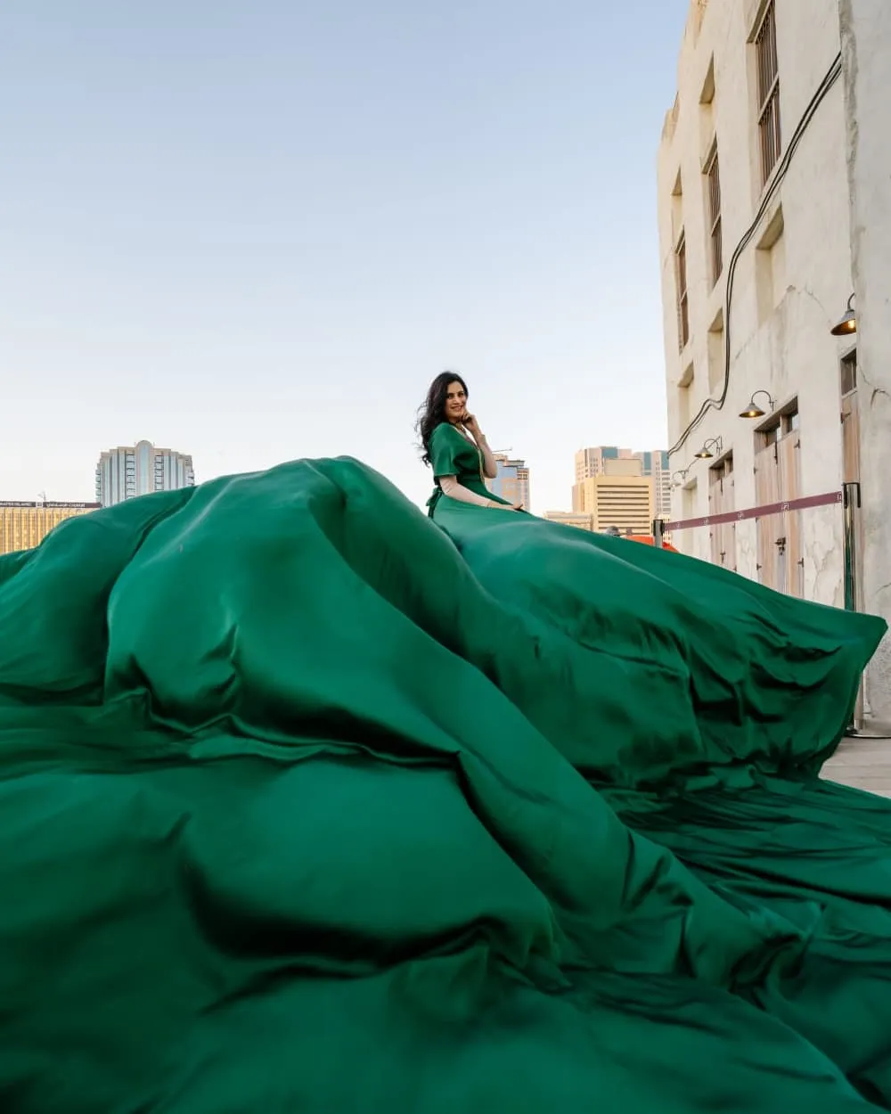 Woman in a very long dark-green gown spread across the ground in front of city buildings