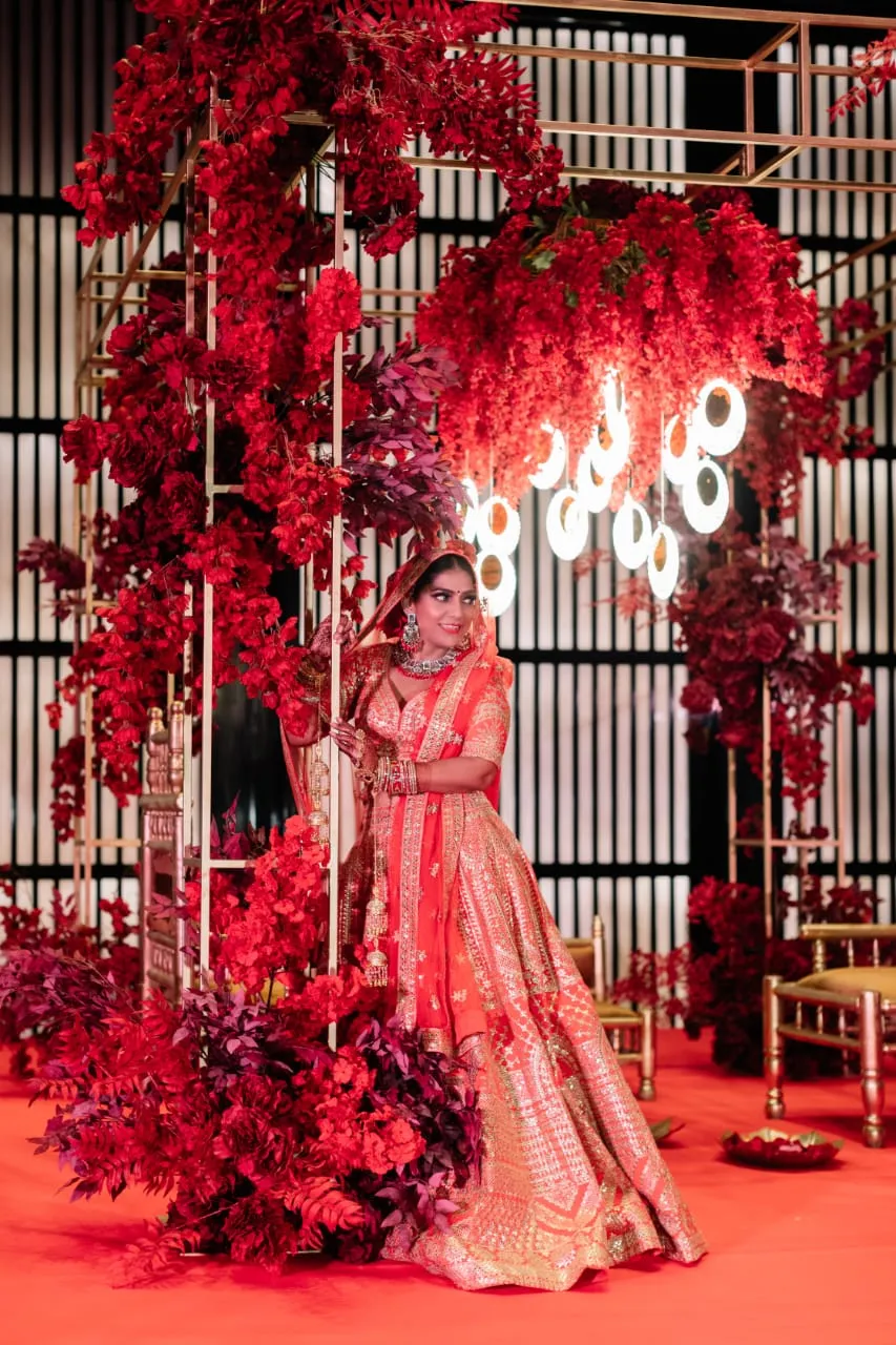 Woman in a red and gold lehenga standing under red floral arrangements on a red-lit set
