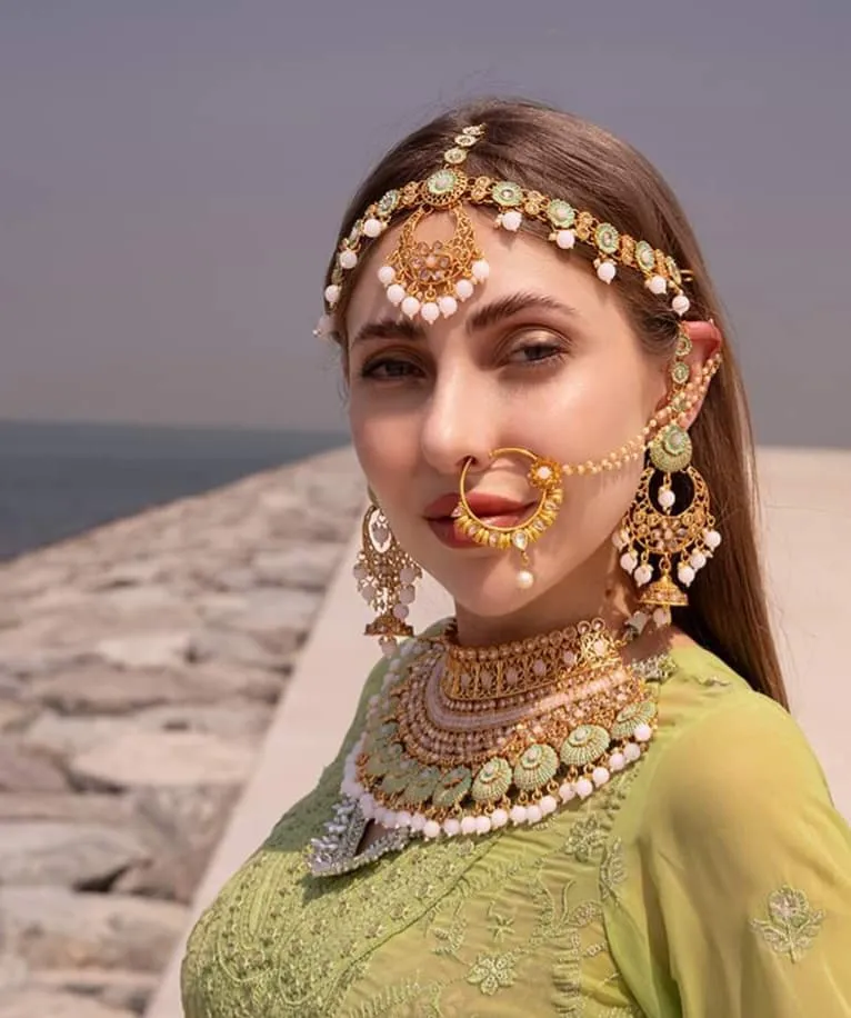 Woman in an olive-green outfit wearing a gold nose chain, forehead ornament and layered necklaces by the waterfront