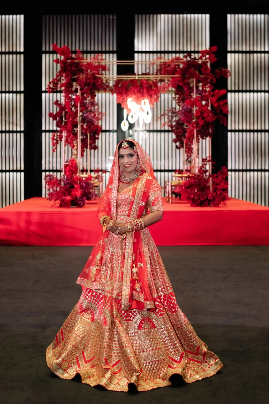 Bride in a red and gold lehenga with a veil standing on a stage dressed with red flowers