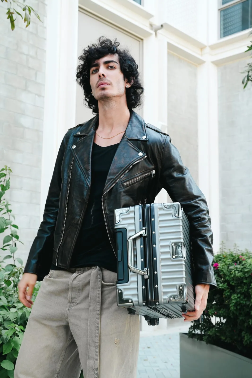 Young man in a black leather jacket holding a silver suitcase in front of a pale building