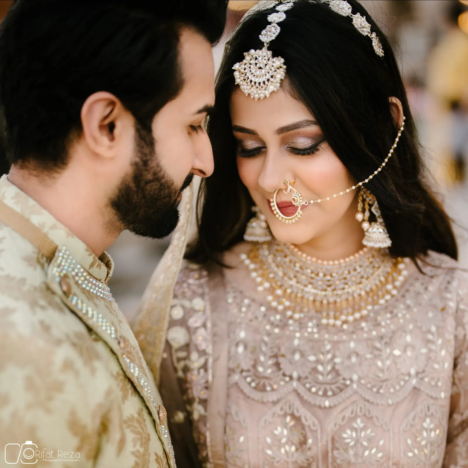 Couple in wedding outfits, the woman in a pink embellished outfit with a forehead ornament and nose chain, the man in a beige kurta