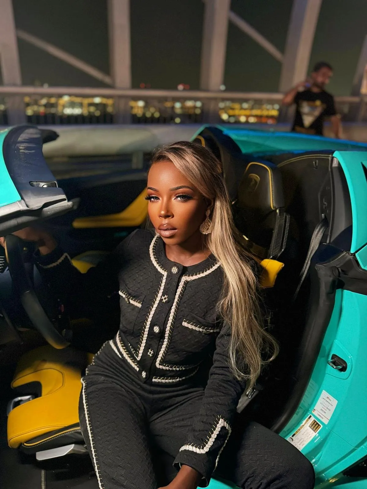 Woman with long blonde hair in a black cardigan sitting in a turquoise sports car